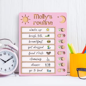 May include: A pink wooden chart with a daily routine for a child named Molly. The chart has a sun and moon icon at the top and includes activities like wake up, brush teeth, breakfast, get dressed, feed Milo, cleaning, dinner, and go to bed. Each activity has a check mark and an X next to it.