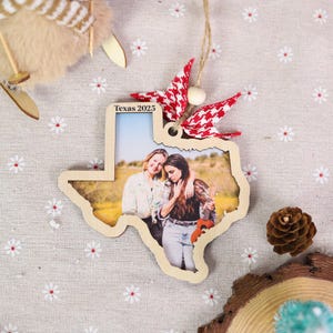 May include: Wooden Texas-shaped ornament with a photo of two people. The ornament has a red and white patterned bow and the text "Texas 2025". A string is attached for hanging. The ornament is on a floral patterned surface.