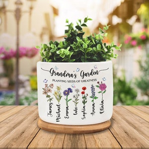 May include: White ceramic planter with a bamboo base. The planter has a floral design with butterflies and the text "Grandma's Garden Planting Seeds of Greatness". The names James, Michael, Emma, Luke, Sophia, Anna, and Olivia are written on the planter.