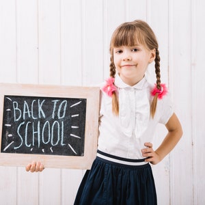May include: A young girl with pigtails wearing a white blouse and a navy blue skirt holds a chalkboard with the words "Back to School" written in white chalk.
