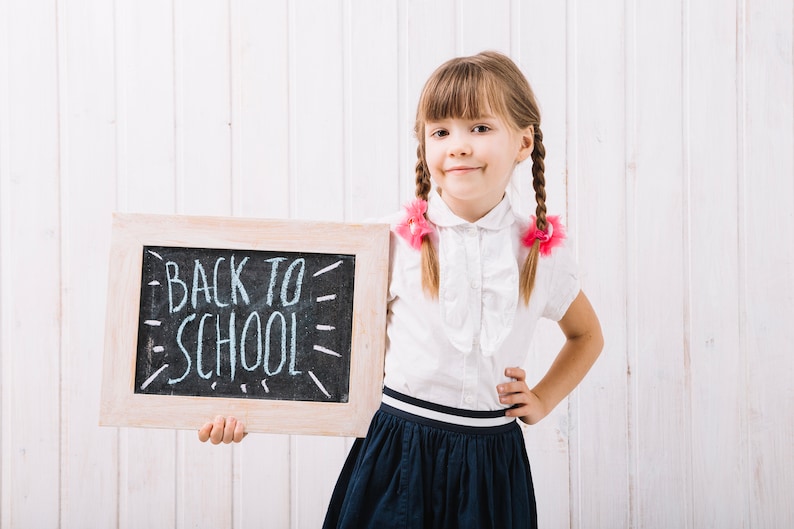 May include: A young girl with pigtails wearing a white blouse and a navy blue skirt holds a chalkboard with the words "Back to School" written in white chalk.