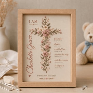 Może przedstawiać: Oprawiona haftowana grafika przedstawiająca kwiatowy krzyż w odcieniach r&oacute;żu i zieleni. Tekst zawiera słowa I AM, Charlotte Grace oraz wersety biblijne, takie jak beautiful, chosen, a child of God i BAPTIZED IN JUNE 2026.