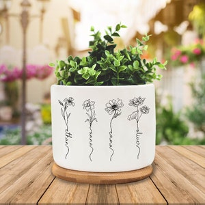 May include: White ceramic planter with a bamboo base. The planter has four black line drawings of flowers with the names "Ethan", "Ariana", "Emma", and "Oliver" written underneath each flower.