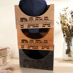 May include: A wooden hat rack with three tiers, each featuring a cutout for a baseball cap. The top tier has a blue baseball cap in the cutout. Each tier is engraved with the word "PAPA" in large letters with an American flag design and the names "Hilary, Beatrix, Larissa" underneath.