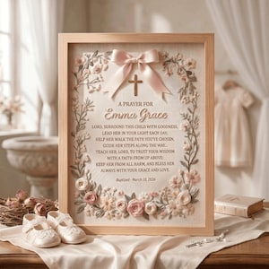 May include: A framed wall art piece featuring a prayer for Emma Grace, dated March 10, 2026. The design includes a central wooden cross, a light pink satin ribbon bow, and a border of embroidered pink and white flowers on a cream fabric background.