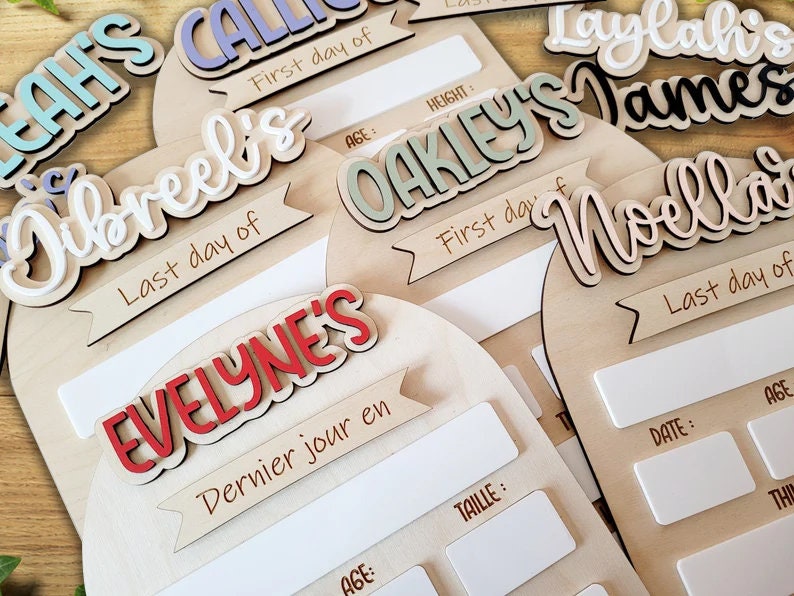May include: Set of six wooden signs with white, black, and red lettering. Each sign has a name, "Jibreel's", "Oakley's", "Noella", "Evelyn's", "Laylah's", and "James", and a banner with the text "First day of" or "Last day of".