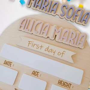May include: A wooden growth chart with a light brown background. The chart has a white banner with the text "First day of" and spaces to record the date, age, height, and things the child loves. The chart is decorated with two wooden name plaques, one with the names "Maria Sofia" in purple letters and the other with the names "Alicia Maria" in pink letters.