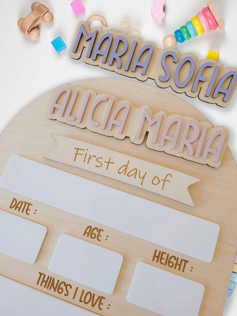 May include: A wooden growth chart with a light brown background. The chart has a white banner with the text "First day of" and spaces to record the date, age, height, and things the child loves. The chart is decorated with two wooden name plaques, one with the names "Maria Sofia" in purple letters and the other with the names "Alicia Maria" in pink letters.