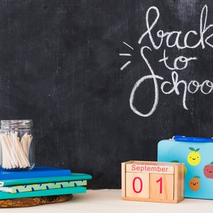 May include: A chalkboard with the words "back to school" written in white chalk. There is a stack of notebooks, a jar of pencils, a lunchbox with a fruit design, and a wooden calendar block that says "September 01".