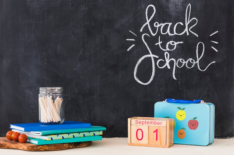 May include: A chalkboard with the words "back to school" written in white chalk. There is a stack of notebooks, a jar of pencils, a lunchbox with a fruit design, and a wooden calendar block that says "September 01".
