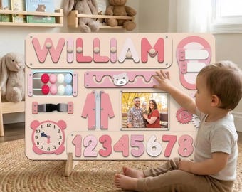 May include: A pink and white wooden activity board with the name "WILLIAM" in large letters. The board features various interactive elements, including a clock, numbers, and a photo frame. A child is interacting with the board.