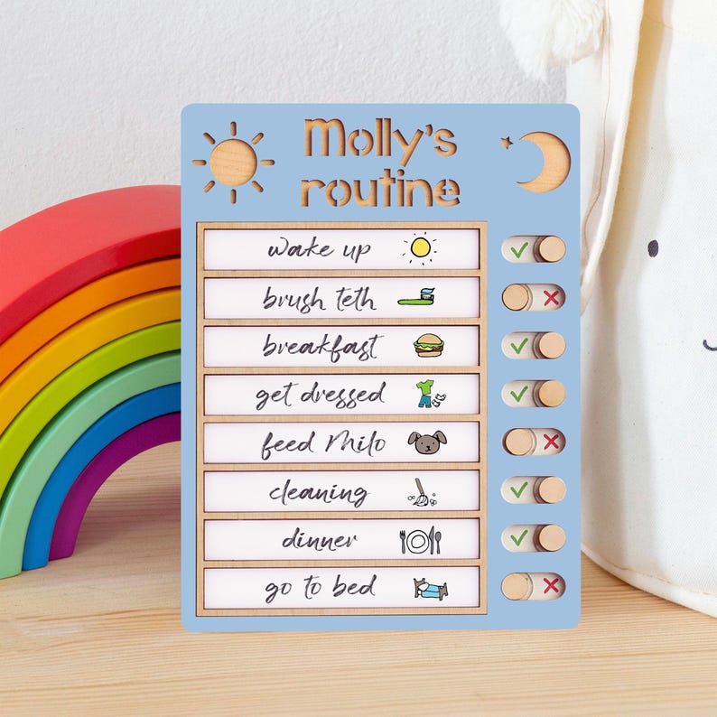 Puede incluir: Un cuadro de rutina diaria de madera azul claro para Molly, con un dise&ntilde;o de sol y luna. El cuadro incluye tareas como "despertarse", "cepillarse los dientes" y "acostarse", con casillas de verificaci&oacute;n para marcar la finalizaci&oacute;n. Un juguete de arco&iacute;ris colorido est&aacute; en el fondo.