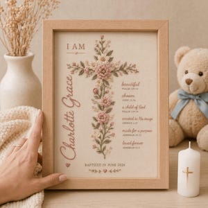 Może przedstawiać: Oprawiona w ramkę haftowana praca przedstawiająca motyw kwiatowego krzyża w odcieniach r&oacute;żu i zieleni. Tekst zawiera słowa I AM, Charlotte Grace, wersety religijne oraz datę BAPTIZED IN JUNE 2026 na jasnym materiale.