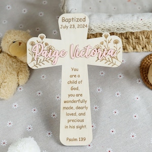 May include: A wooden cross with the name "Paige Victoria" in pink script, adorned with floral accents. The cross includes the text "Baptized July 23, 2024" and a religious verse from Psalm 139. The cross is a light beige color.
