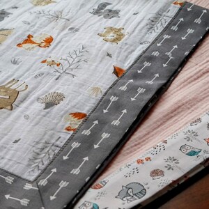 May include: Two baby blankets with a woodland animal theme. One blanket is white with gray arrows around the border and features a variety of animals including foxes, deer, and owls. The other blanket is pink with a white border and features a variety of animals including foxes, owls, and hedgehogs.
