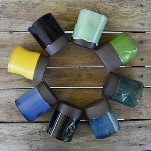 Peut inclure: Ensemble de neuf mugs en c&eacute;ramique avec une bordure brune et des gla&ccedil;ures de diff&eacute;rentes couleurs. Les mugs sont dispos&eacute;s en cercle sur une surface en bois.
