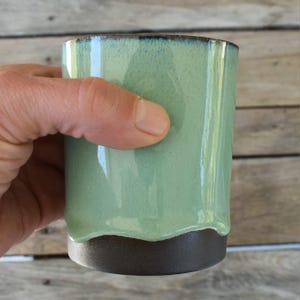 Peut inclure: Une tasse en c&eacute;ramique verte avec un bord et une base brun fonc&eacute;. La tasse a un bord l&eacute;g&egrave;rement irr&eacute;gulier et ondul&eacute;.