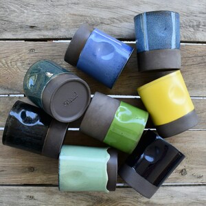 Peut inclure: Un ensemble de huit tasses en c&eacute;ramique de diff&eacute;rentes couleurs, dont le bleu, le vert, le jaune et le noir. Les tasses ont un gla&ccedil;age brun sur la moiti&eacute; inf&eacute;rieure et une vari&eacute;t&eacute; de gla&ccedil;ages sur la moiti&eacute; sup&eacute;rieure. Les tasses sont dispos&eacute;es en cercle sur une surface en bois.