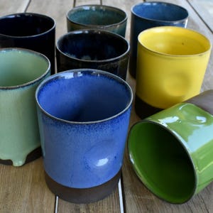 Peut inclure: Une collection de huit tasses en c&eacute;ramique de diff&eacute;rentes couleurs, notamment vert, bleu, jaune et marron. Les tasses sont toutes de formes et de tailles diff&eacute;rentes et sont dispos&eacute;es en rang&eacute;e sur une surface en bois.