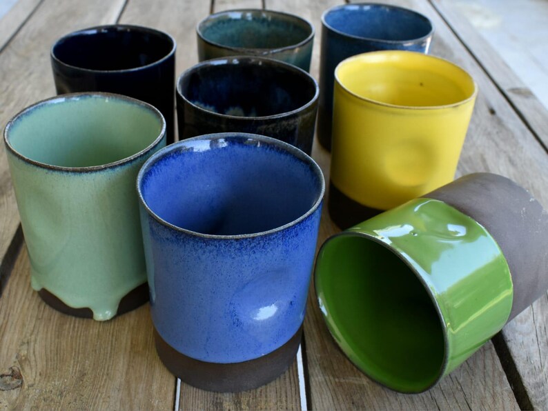 Peut inclure: Une collection de huit tasses en c&eacute;ramique de diff&eacute;rentes couleurs, notamment vert, bleu, jaune et marron. Les tasses sont toutes de formes et de tailles diff&eacute;rentes et sont dispos&eacute;es en rang&eacute;e sur une surface en bois.