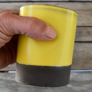 Peut inclure: Une tasse en c&eacute;ramique jaune avec une bande noire autour du bas. La tasse est en poterie et a une finition lisse et brillante.