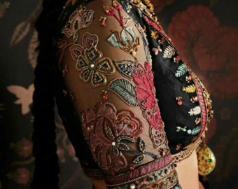 Puede incluir: Una blusa negra transparente con intrincados bordados florales y adornos de cuentas. Las mangas son de tres cuartos y presentan múltiples capas de ribetes de colores.