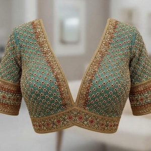 Op de afbeelding: Een groenblauwe blouse met een diepe V-hals en korte mouwen, versierd met een ingewikkeld gouden rasterborduurpatroon. Het ontwerp bevat kleine rode en witte bloemaccenten en een brede, rijkelijk versierde gouden rand.