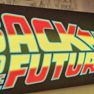 Op de afbeelding: Een rechthoekig bord met de tekst "BACK TO THE FUTURE" in rode en gele letters. Het bord heeft een zwarte achtergrond en een lichtblauwe omtrek. Het bord is licht gebogen en heeft een retro-ontwerp.