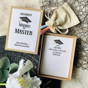 Könnte beinhalten: Zwei rechteckige Schachteln aus Kraftpapier mit weißen Karten. Eine Karte zeigt den Text GESCHAFFT! MATHEO IST MASTER Herzlichen Glückwunsch! mit Doktorhut-Symbol. Die andere Karte trägt den Text Von HERZEN alles Liebe und Gute zu Deinem ERFOLG! mit Doktorhut. Ein kleiner beiger Beutel mit Kordelzug enthält gerollte Zettel.