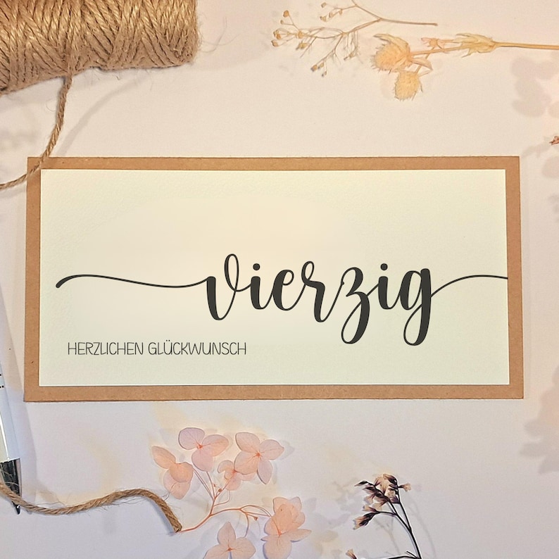 May include: A rectangular greeting card with a cream-colored center and a brown border. The card features the word "Vierzig" in elegant black script, with "HERZLICHEN GL&Uuml;CKWUNSCH" below. Decorative twine and dried flowers are arranged around the card.
