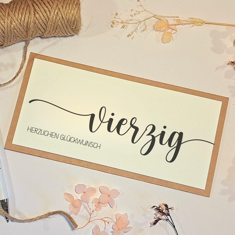 May include: A greeting card with the word "Vierzig" in elegant black script, and "HERZLICHEN GL&Uuml;CKWUNSCH" below. The card has a cream-colored center with a brown border. Twine and dried flowers are arranged around the card.
