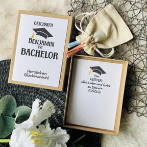 Könnte beinhalten: Zwei rechteckige Pappschachteln mit weißen Karten, die schwarze Doktorhut-Symbole und deutschen Text zeigen. Eine Karte lautet GESCHAFFT! BENJAMIN IST BACHELOR. Die andere lautet Von HERZEN alles Liebe und Gute zu Deinem ERFOLG! Ein kleiner beiger Kordelzugbeutel liegt bei.