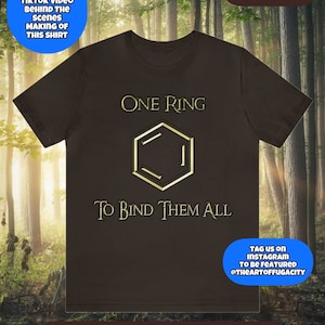 Peut inclure: Un t-shirt marron avec un motif hexagonal doré sur lequel est imprimé le texte "One Ring" et "To Bind Them All". Le t-shirt est présenté sur un fond flou d'arbres verts.