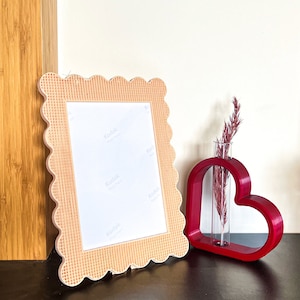 Könnte beinhalten: Ein gewellter, hellbrauner Bilderrahmen mit einem weißen Passepartout und einem leeren weißen Einsatz. Eine rote herzförmige Vase mit einem durchsichtigen Glasrohr enthält einen Trockenblumenstrauß.