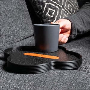May include: A black, multi-layered, heart-shaped tray with an orange stripe sits on a grey sofa. A grey mug sits on the tray.