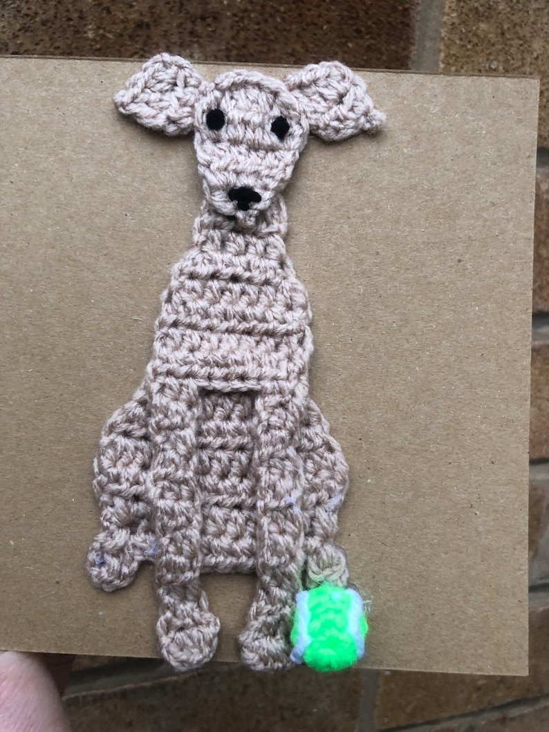 May include: A crocheted brown dog with black eyes and a black nose, holding a green tennis ball.