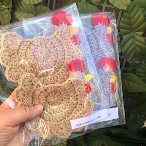 May include: Two sets of crochet chicken coasters packaged in clear plastic sleeves. One set features beige yarn, while the other set features light blue yarn. Each coaster has a red comb and yellow beak. A white label reads Chicken coasters x 2 - beige.