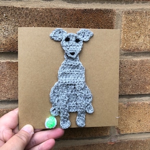 May include: A gray crocheted dog sitting on a brown card with a green ball at its feet.