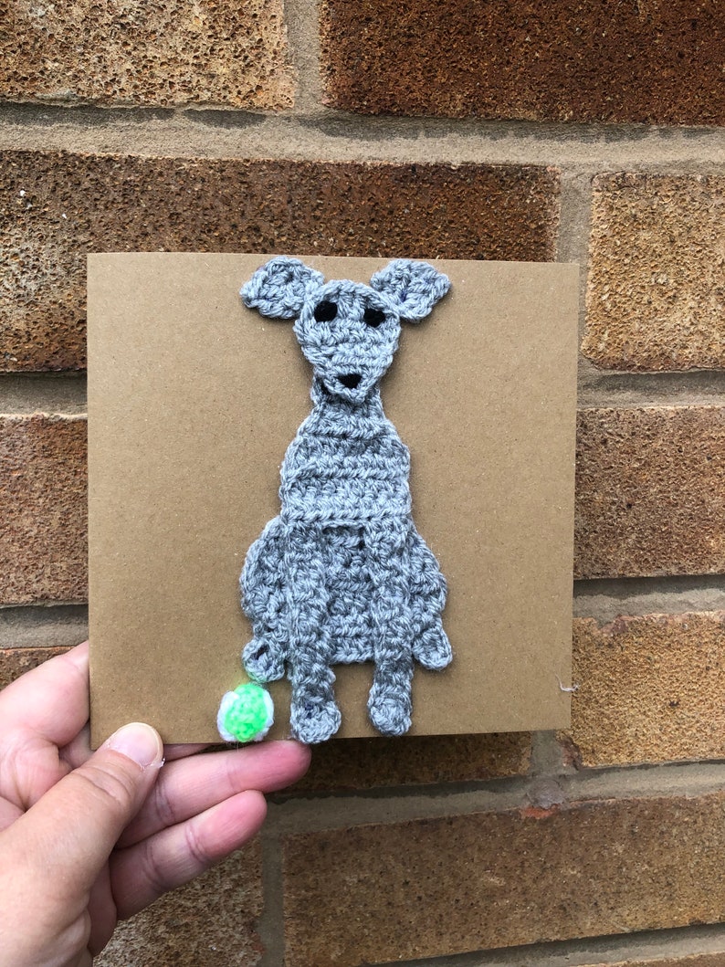 May include: A gray crocheted dog sitting on a brown card with a green ball at its feet.