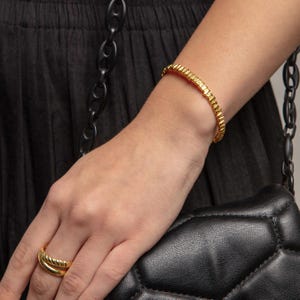 Könnte beinhalten: Ein goldfarbener Metallarmreif mit einer texturierten, geriffelten Oberfläche und ein passender goldfarbener Ring mit einem ähnlichen gedrehten, seilartigen Design. Beide Schmuckstücke weisen eine polierte, reflektierende Oberfläche und einen einheitlichen metallisch-gelben Farbton auf.
