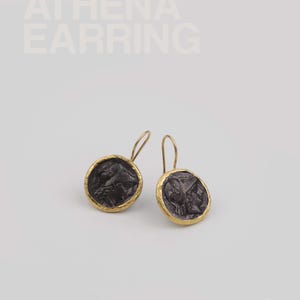 May include: A pair of Athena earrings featuring a dark, textured coin design set in gold-coloured metal. The earrings have a hook closure and are displayed on a white background. The text "ATHENA EARRING" is at the top.