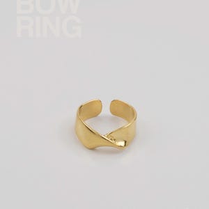 Könnte beinhalten: Ein goldfarbener Ring mit einem verdrehten Design. Der Ring ist hinten offen und trägt den Text "BOW RING" in der oberen linken Ecke. Der Ring ist vor einem weißen Hintergrund platziert.