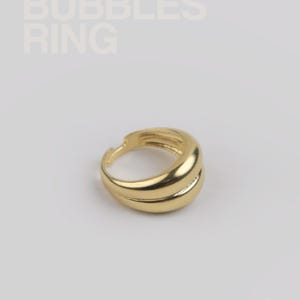 Könnte beinhalten: Ein goldfarbener Ring mit Doppelband-Design. Der Ring hat eine glatte, polierte Oberfläche und ist vor einem weißen Hintergrund platziert. Der Text "BUBBLES RING" ist oben links zu sehen.