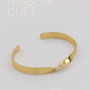 Könnte beinhalten: Ein goldfarbenes Armband im Ribbon-Cuff-Stil. Das Armband hat ein verdrehtes Design in der Mitte und ist an den Enden offen. Der Text "RIBBON CUFF" befindet sich in der oberen linken Ecke.