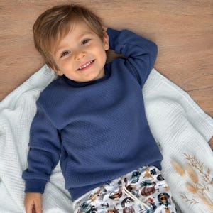 Könnte beinhalten: Ein langärmeliger, marineblauer Pullover aus Waffelstrick mit Rundhalsausschnitt und gerippten Bündchen, kombiniert mit einer weißen Hose, die mit einem bunten Muster aus verschiedenen Tieren und nautischen Motiven bedruckt ist.