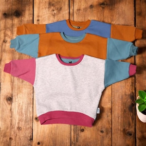 Op de afbeelding: Drie shirts met lange mouwen met contrasterende kleur mouwen en halslijnen. De shirts zijn grijs, oranje en blauw met teal mouwen en halslijnen. De shirts liggen op een houten oppervlak.