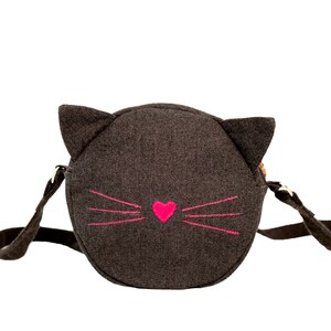 Puede incluir: Un bolso bandolera de fieltro gris en forma de cara de gato con bigotes bordados en rosa y un coraz&oacute;n rosa.
