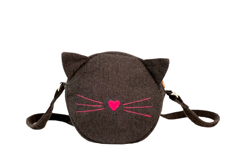 Puede incluir: Un bolso bandolera de fieltro gris en forma de cara de gato con bigotes bordados en rosa y un coraz&oacute;n rosa.