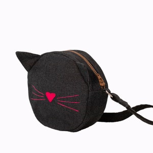 Puede incluir: Un peque&ntilde;o bolso bandolera de mezclilla negro con un dise&ntilde;o de cara de gato. El gato tiene una nariz de coraz&oacute;n rosa y bigotes rosas.