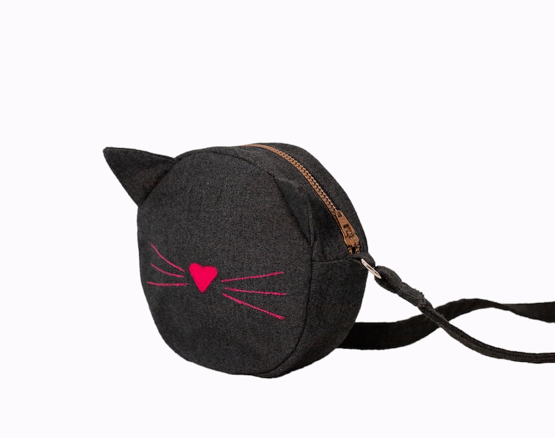 Puede incluir: Un peque&ntilde;o bolso bandolera de mezclilla negro con un dise&ntilde;o de cara de gato. El gato tiene una nariz de coraz&oacute;n rosa y bigotes rosas.